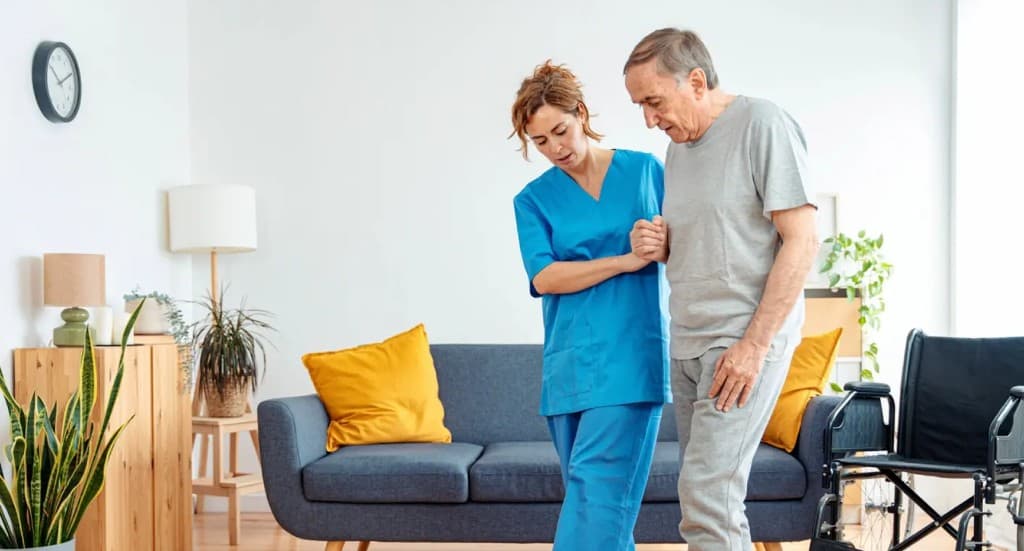 Caregiver in blue scrubs assisting an elderly man in a bright, modern living room