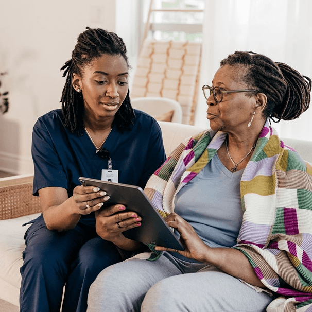 Caregiver in blue scrubs with an older woman on a sofa in a bright living room