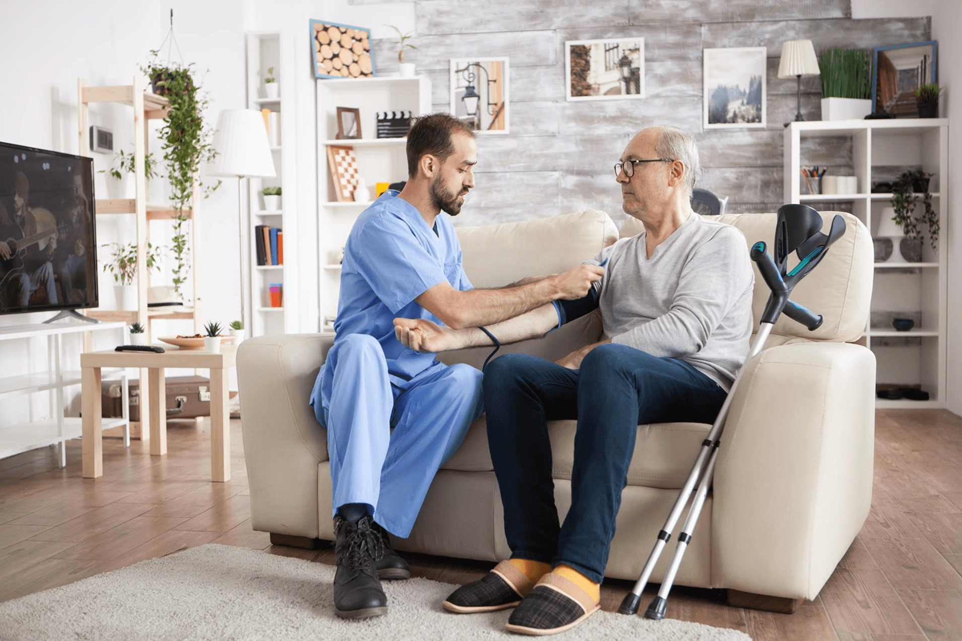 Caregiver in light blue scrubs with a glass of water, smiling at an elderly woman in bed in a warm bedroom setting