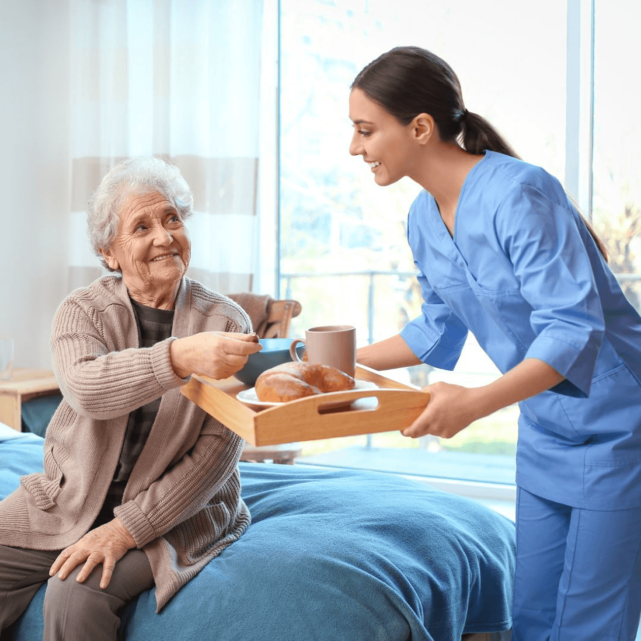 Caregiver in light blue scrubs with stethoscope gently placing her hand on an elderly woman's shoulder in a calm indoor setting