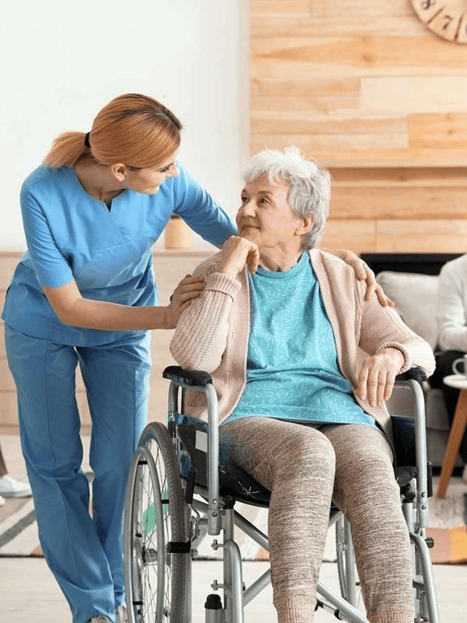 Caregiver in pink scrubs kneeling and holding hands with an elderly woman seated on a sofa in a living room